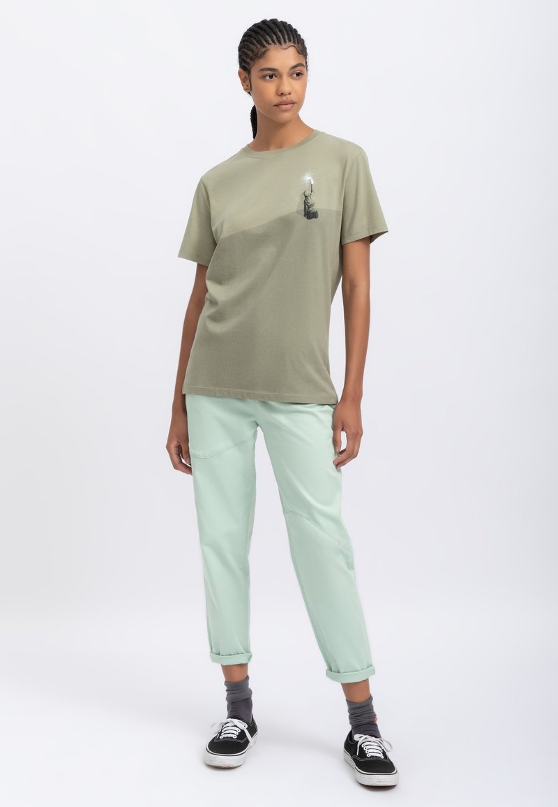 T-shirt verde oliva a maniche corte con grafica centrale di una figura, abbinato a pantaloni mint green arrotolati e sneakers nere.