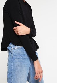 Maglione nero a maglia con scollatura rotonda e orlo a strati. Abbinato a jeans chiari strappati, che mettono in mostra un look casual.