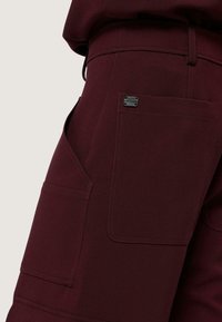 Bordeaux kleurige broek gemaakt van een getextureerde stof, met meerdere zakken en een klein merklabel op de achterkant.