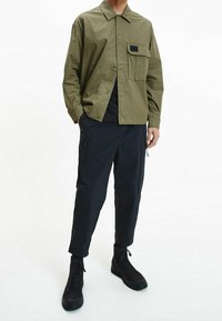 Olivengrüne, leichte Jacke mit einer Fronttasche und schwarzen Akzenten, kombiniert mit dunklen, lockeren, strukturierten Hosen und schwarzen Stiefeln.