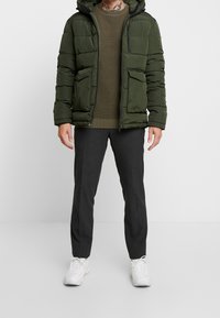 Hombre con chaqueta acolchada verde oscuro, suéter verde oliva, pantalones gris oscuro y zapatillas blancas de pie sobre un fondo liso.