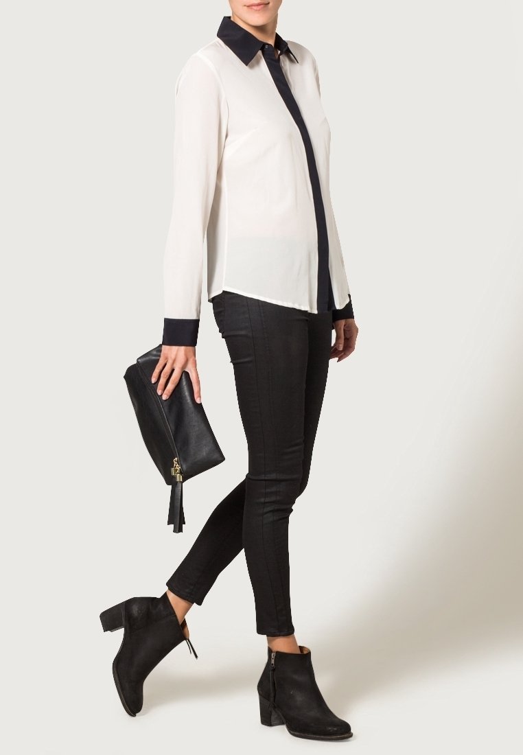 Langärmliges weißes Hemd mit einem marineblauen Kragen, kombiniert mit schwarzen Skinny-Jeans und schwarzen Stiefeletten; hält eine schwarze Clutch mit einem Quasten.