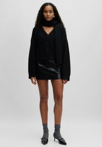 Zwarte knoopsluiting cropped cardigan, zwarte mini rok van faux leer met krokodillenpatroon, grijze gebreide sokken en puntige zwarte hakken.