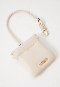 Petit sac en cuir beige avec une poignée en boucle et un clip doré, présentant le lettrage doré "GUESS" sur le devant.