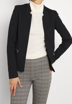 Vrouw draagt een zwarte blazer met ritszakken, een crèmekleurige coltrui en grijze geruite broek, staat met één hand op de heup en de andere ontspannen.