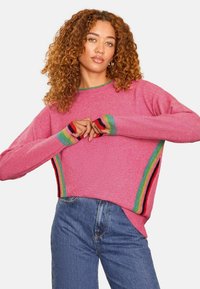 Ružový pletený sveter s voľným strihom a viacfarebnými pruhmi na manžetách a bočnom šve, v kombinácii s modrými džínami.