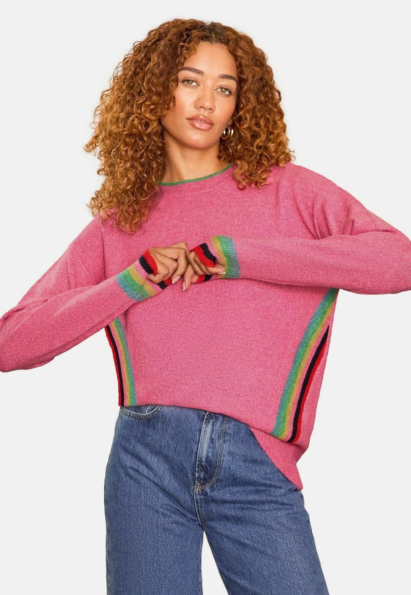 Ružový pletený sveter s voľným strihom a viacfarebnými pruhmi na manžetách a bočnom šve, v kombinácii s modrými džínami.