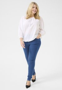 Blouse blanche à pois, à manches longues, assortie à un jean skinny bleu et à des escarpins noirs pointus. La blouse a une coupe décontractée et des poignets élastiques.
