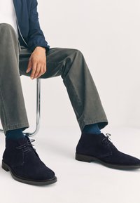 Marineblaue Chukka-Stiefel aus Wildleder mit abgerundetem Zehenbereich, Schnürdesign und glatter schwarzer Sohle. Getragen mit olivgrünen Hosen und blauen Socken.