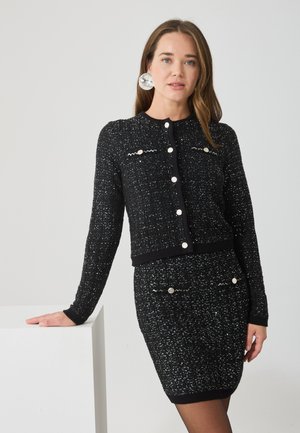 Čierny pletený kardigán a sukňový set s textúrovanou tkaninou, okrúhlymi striebornými gombíkmi a kontrastnými bielymi detailmi na vreckách.