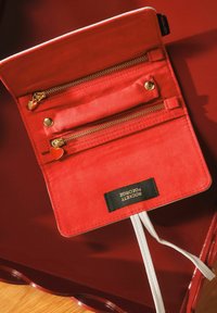 Portefeuille en daim rouge avec des zips dorés, deux poches zippées, un détail en forme de cœur et un cordon blanc. L'étiquette indique "ROCKETT SEGEORGE."