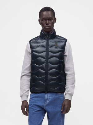 Mann trägt dunkelblaue gesteppte ärmellose Weste über hellgrauem Langarmshirt und blauer Jeans vor weißem Hintergrund.