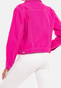 Fuchsia denimjakke med kontrastsyning, kort design og knappedetalje ved kanten. Stylet med hvide ribstrikkede bukser.