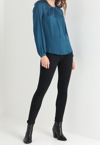 Teal långärmad blus med rundad halsringning och plisserade detaljer, parats med svarta skinny jeans och svarta ankelstövlar.