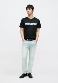 Μαύρο βαμβακερό t-shirt με τη φράση "DON DUP" σε μπλε γράμματα, ανοιχτόχρωμο μπλε τζιν, μαύρη ζώνη και μαύρα μοκασίνια. Το μοντέλο στέκεται με τα χέρια στις τσέπες.