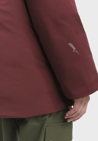 Bordeaux waterdichte jas met lange mouwen, een gladde textuur en een klein zilveren accentlogo op de mouw. Groene broek gedeeltelijk zichtbaar.