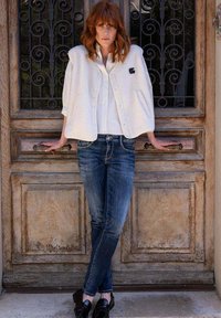 Veste en polaire blanche avec une surface texturée, portée par-dessus une chemise à boutons, associée à un jean bleu et des chaussures noires, se découpant sur une porte en bois.