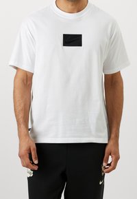 Bílá bavlněná tričko s krátkým rukávem a černým obdélníkovým nášivkou loga Nike umístěnou na hrudi. Design s kulatým výstřihem.