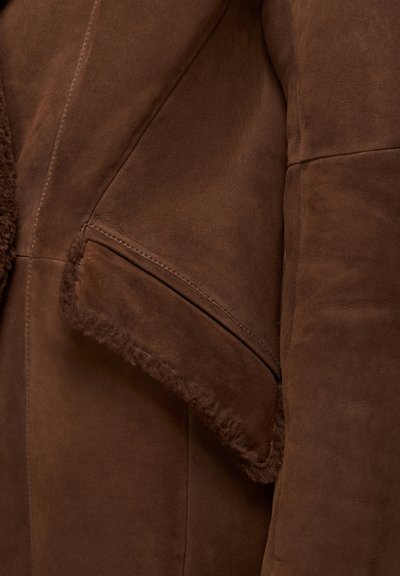 Pruun seemisnahkse jope pehme shearling-kaelusega ja küljetaskuga, millel on nähtavad õmblused. Sile tekstuur, keha vormi järgiv lõige.