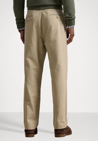 Beige pamut nadrág laza szabással, dupla hátsó zsebekkel és egy kis logóval. Az anyag sima tapintású.