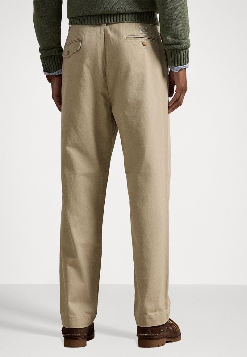 Pantaloni di cotone beige con vestibilità rilassata, dotati di doppie tasche posteriori e una piccola etichetta con logo. Il tessuto ha una texture liscia.