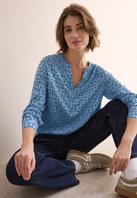 Camicia a maniche lunghe azzurro chiaro con un motivo astratto nero, patta con bottoni e polsini arrotolati, abbinata a jeans scuri e sneakers grigie.