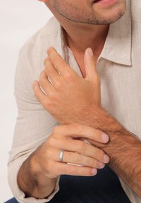 Silver metal ring on a hand, featuring a smooth circular band. The hand is lightly tanned with short nails. Light-colored linen shirt visible.