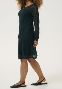 Lace dress in dark green, long-sleeved, with a flared hem. Worn with black slingback shoes featuring circular accents.