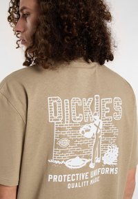 Beige cotton t-shirt featuring a white graphic design on the back, showcasing a figure against a brick wall with text reading "DICKIES."