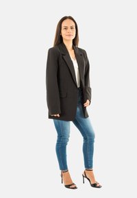 Blazer noir à manches longues, porté sur un top blanc, associé à un jean skinny bleu et des escarpins noirs à brides. Fond uni.