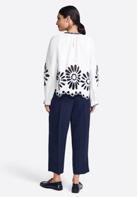 Blusa bianca con ricami floreali neri, maniche a campana e orlo a scallop. Pantaloni blu navy. Materiale leggero, vestibilità ampia, dettaglio di chiusura posteriore.