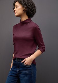 Bordeaux coltrui met lange mouwen, gladde textuur, gecombineerd met donkerblauwe denim jeans. Model staat tegen een grijze achtergrond.