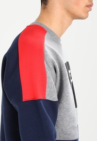 Grå, marinblå och röd färgblockerad sweatshirt med rund hals, mjuk textur och framträdande logotyp på framsidan. Har ribbade ärmslut.