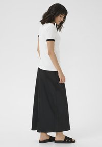 Chemise polo blanche avec des poignets noirs, associée à une longue jupe noire et des sandales noires. La tenue présente une coupe décontractée et des textures lisses.