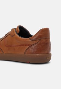 Hnědé kožené tenisky s texturovaným povrchem, prošívanými detaily a gumovou podrážkou s malým červeným prvkem. Nízkoprofilový design.
