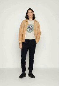 Veste beige sur un t-shirt graphique blanc, pantalons de survêtement noirs et baskets noires. La veste a une texture lisse ; le t-shirt présente un motif circulaire.