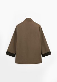 Cappotto leggero marrone con una vestibilità rilassata, dotato di maniche a tre quarti e polsini neri texture. Design minimalista senza chiusure visibili.