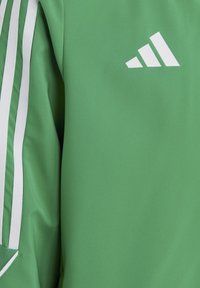 Grüne Sportjacke mit weißem Adidas-Logo, strukturiertem Stoff und drei weißen Streifen entlang der Schultern. Modernes, tailliertes Design.