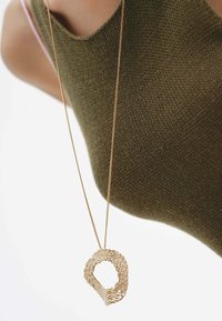 Collar de oro con un colgante en espiral texturizado, suspendido de una cadena fina, llevado sobre un top de punto verde oliva.