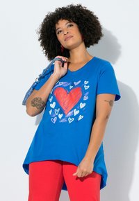 Femme aux cheveux bouclés portant un t-shirt bleu avec un grand motif de cœur rouge et un pantalon rouge, tenant une sangle de sac à main tressée bleue.
