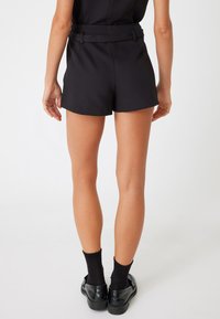 Pantalones cortos negros hechos de una tela suave, con una cintura ajustada que incluye un cinturón a juego, y un dobladillo que cae a mitad del muslo. Zapatos negros con calcetines texturizados.