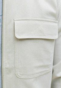 Nærbilde av en teksturert hvit jakkelomme med klaff og synlige sømmer, ved siden av en vertikal sølvglidelås.