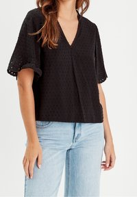 Haut noir en tissu texturé avec un décolleté en V et des manches courtes. Présente des accents en œillets sur les manches et une coupe décontractée. Associe avec un jean bleu clair.