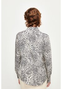 Langärmlige Bluse in Creme mit schwarzen abstrakten Punkten, ausgestattet mit einem hohen Kragen, einer durchgehenden Knopfleiste und einem abgerundeten Saum. Leichter Stoff mit glatter Textur.