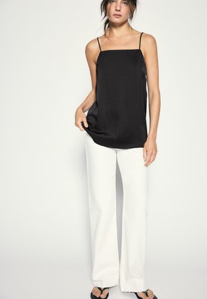 Femme aux cheveux foncés portant un haut noir sans manches, un pantalon blanc ample et des sandales noires à bout ouvert sur un fond clair uni.