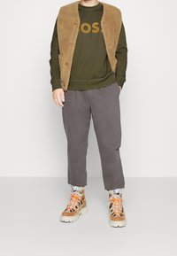 Chaleco de forro polar marrón de gran tamaño sobre un sudadera verde oliva con "BOSS" en dorado. Pantalones grises de corte relajado y zapatillas beige con suela gruesa y cordones naranjas.