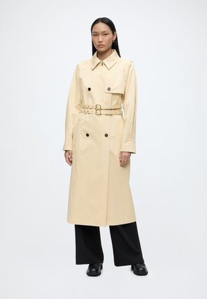 Mujer con pelo largo y oscuro que lleva un abrigo trench beige con cinturón, pantalones negros de pierna ancha y zapatos negros, de pie sobre un fondo blanco liso.