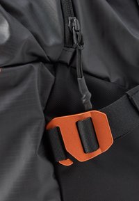 Černý batoh z látky s texturovaným povrchem, s oranžovou plastovou sponou a černým popruhem. Obsahuje detaily se zipem a stahovací šňůrkou.