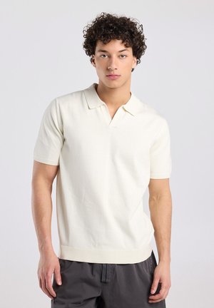 Jonge man met krullend donker haar, gekleed in een wit poloshirt met korte mouwen en donkere broek, tegen een effen lichte achtergrond.
