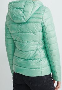 Chaqueta acolchada verde menta con capucha, textura acolchada y costuras horizontales. La tela tiene un acabado brillante. Se llevan pantalones negros debajo.